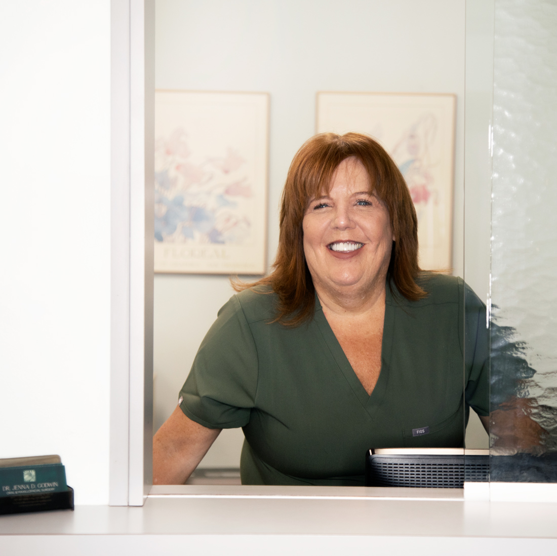 Smiling woman in green scrubs standing behind a reception desk. mobile